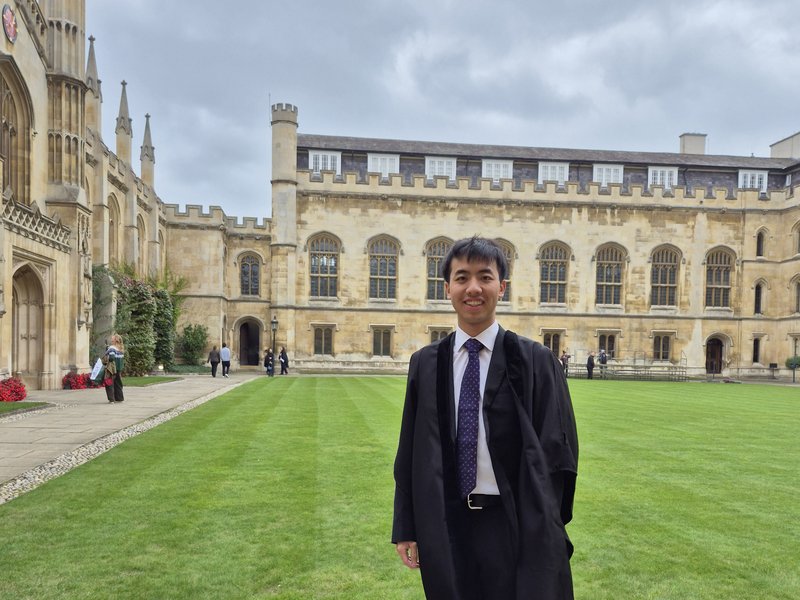 William Yang in suit and matriculation gown standing in the courtyard of Corpus Christi College Cambridge