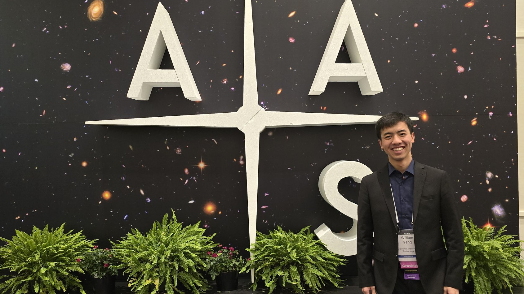 William Yang stands in front of American Astronomical Society conference backdrop smiling at camera.